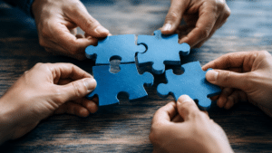 People collaborate to fit blue puzzle pieces together, symbolizing teamwork and problem solving.