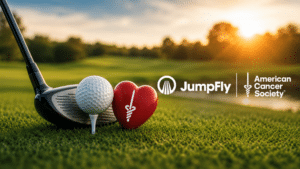 Golf ball on a tee with a club nearby, a red heart-shaped ribbon, and JumpFly + American Cancer Society logos at sunset on a green golf course.