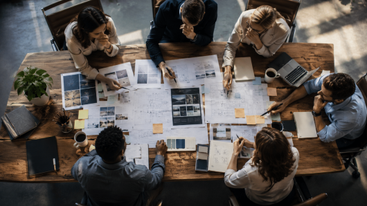 Overhead view of a diverse team around a wooden table, examining blueprints, photos, and sticky notes during a design brainstorm.